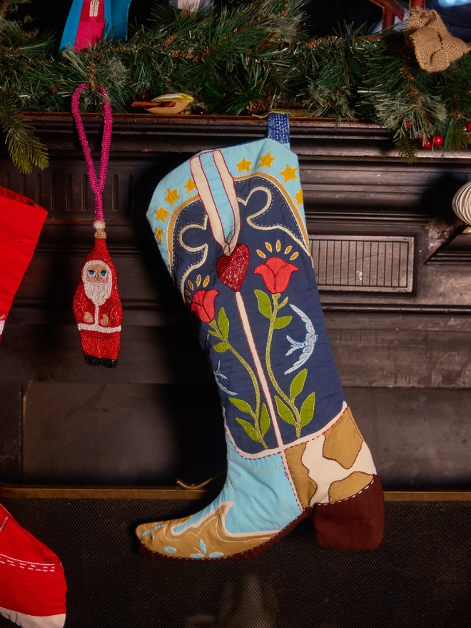 A handcrafted Christmas stocking shaped like a western boot, hanging on a dark fireplace mantel decorated with pine branches and ornaments. The stocking features embroidered floral patterns in red, green, and yellow on a navy blue shaft, with light blue and tan panels near the bottom.