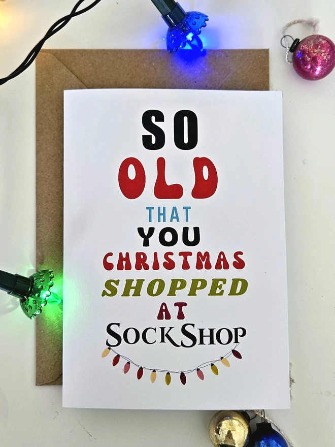 A photograph of the front of a humorous A6 greeting card illustrated using brightly coloured text and the retro shop logo. The text says so old  that you Christmas shopped at Sock Shop with string lights