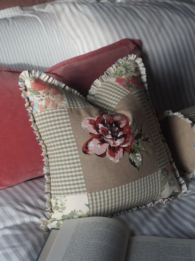 Embroidered floral patchwork cushion in sage gingham and linen, finished with a gathered ruffle trim and styled on a bed.