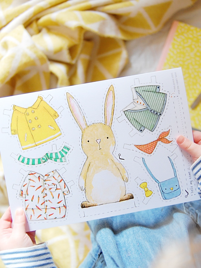 A child is holding a greetings card with an illustrated rabbit paper doll and several outfits on.
