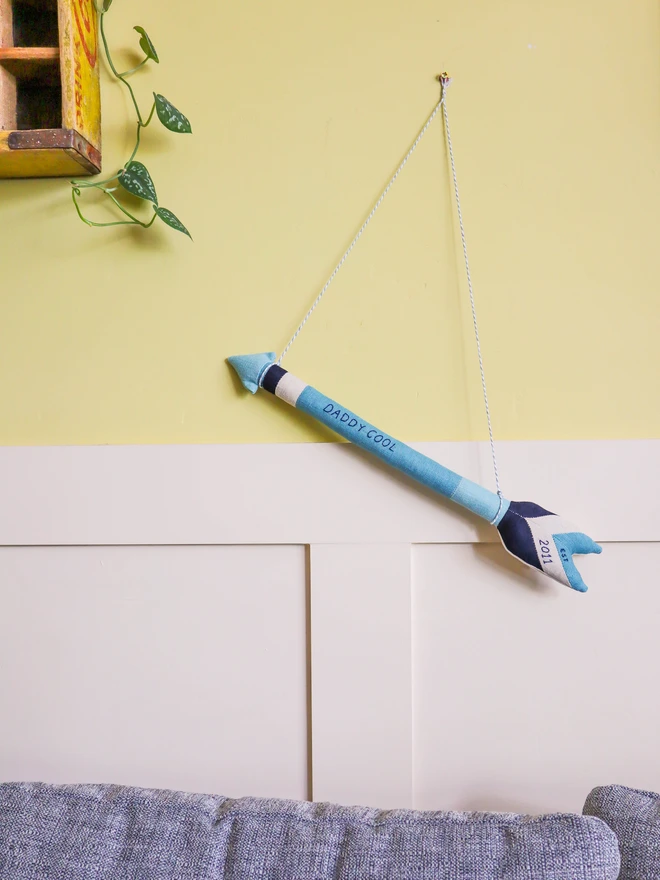 A handmade linen arrow wall hanging in shades of blue and navy, embroidered with the words “DADDY COOL” along the shaft and “2011” on the fletching. A white and grey cord is attached for hanging.