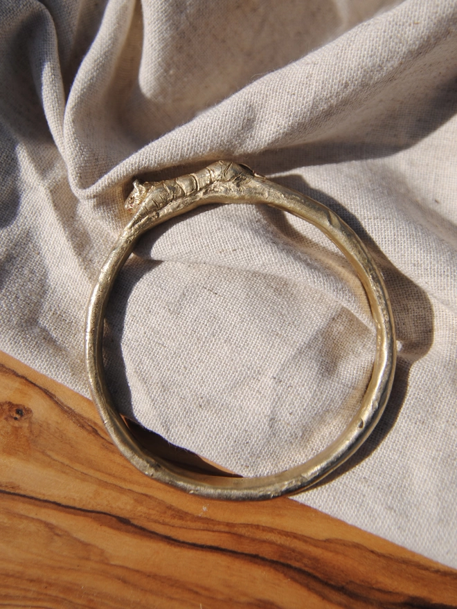 Image of a gold toned brass bangle with a hand carved tiger on the top of the bangle, the bangle is on a piece of raw linen and a piece of rustic wood 