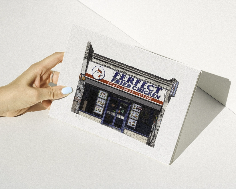 A hand with blue nail polish holding a print featuring an illustration of the Ferfect Fried Chicken shopfront against a white background.