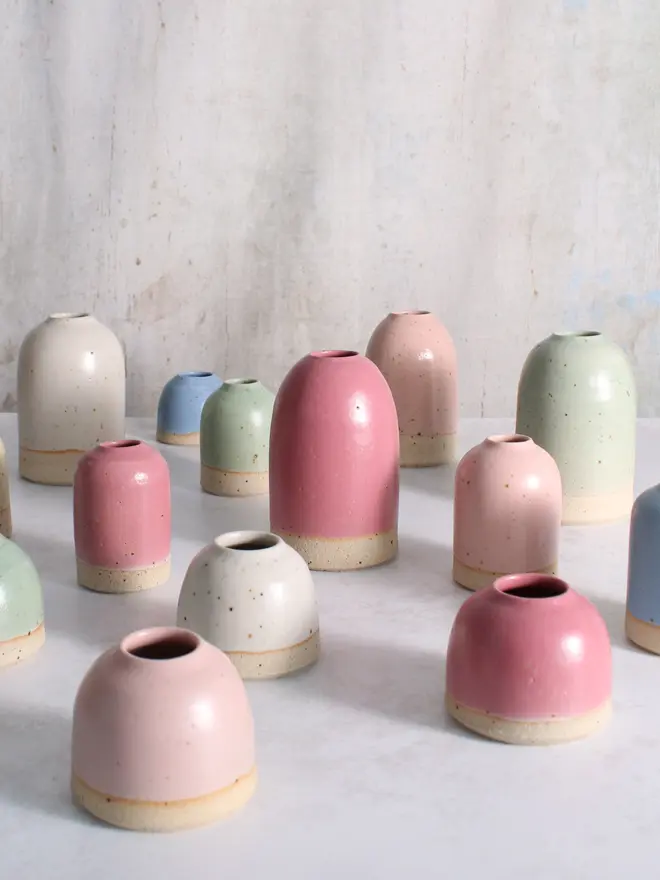 A selection of mini, small and medium ceramic vases in cornflower blue, mint green, pale pink and fuchsia on a white surface against a textured white and grey backdrop. 
