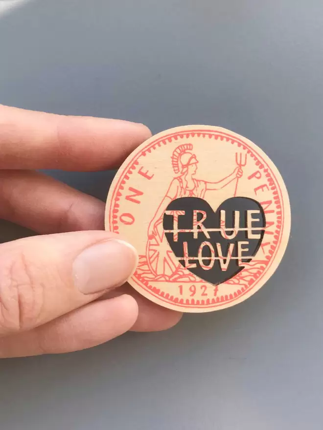 A hand holds the intricate wooden screenprinted Love token coin. The cut out words read 'True Love'