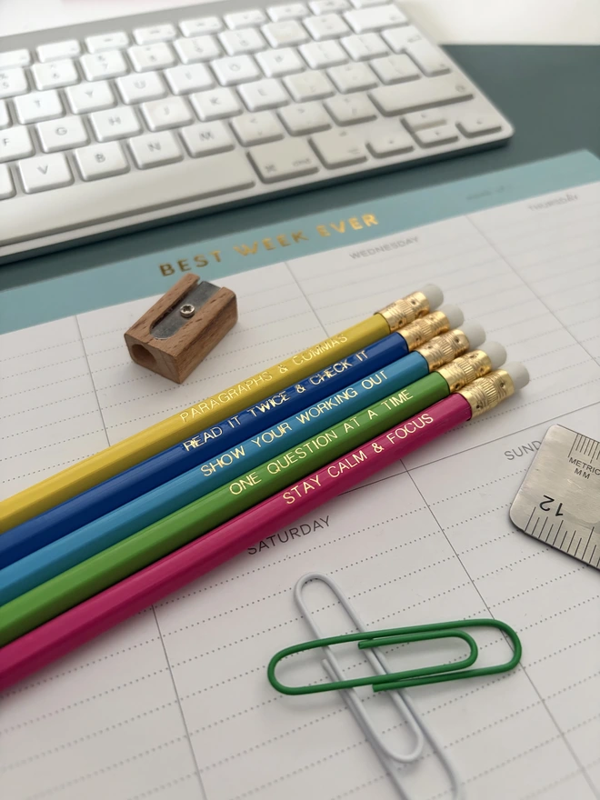 Exam essentials pencil set - Five colourful motivational exam pencils with gold foil lettering are styled on a pastel desk setup with stationery and a weekly planner.