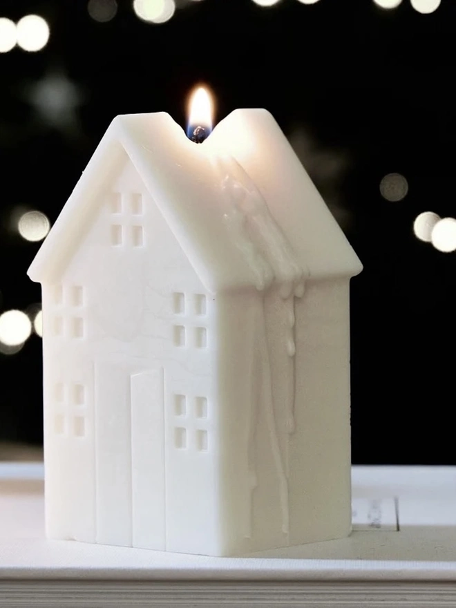 White Christmas Townhouse Candles (Set Of 3). A lit cream house-shaped candle with a pitched roof, embossed windows and a front door, glowing against twinkling bokeh lights. The Scandi-style sculptural candle adds a cosy, nostalgic touch to winter evenings.