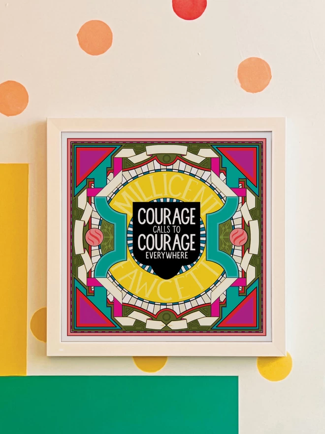 A square illustration with “Courage calls to courage everywhere” written in white against a black background at the centre, surrounded by Millicent Fawcett written in yellow, and bordered with a symmetrical design in white, greens and pinks. It is hung in a white frame on a white wall with yellow, orange, green and blue spots, and a green and yellow rectangle painted in the bottom left hand corner. 