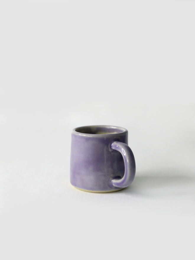 A handmade ceramic espresso mug in amethyst purple glaze, with a chunky loop handle, photographed against a plain light grey background