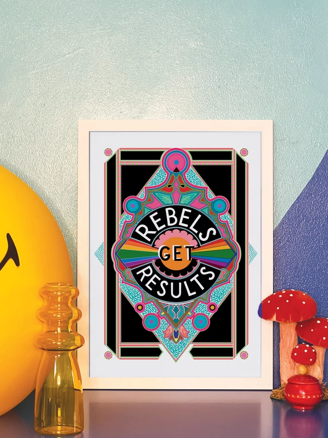 Rebels Get Results is written in white on a black background at the centre of this vibrant, abstract portrait illustration, with a black background and rainbows emitting from the centre, and blue and pink detailing. The picture is in a white frame leaning against a painted blue wall on a blue cabinet. Next to the frame is a large illuminated yellow Smiley lamp, a yellow vase, a model of three red and white toadstools and a small round red wooden pot.