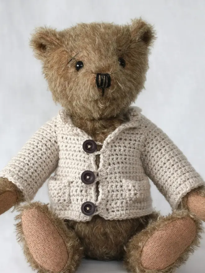 Brown teddy bear sitting upright, wearing a cream knitted cardigan with three buttons, against a plain light background.