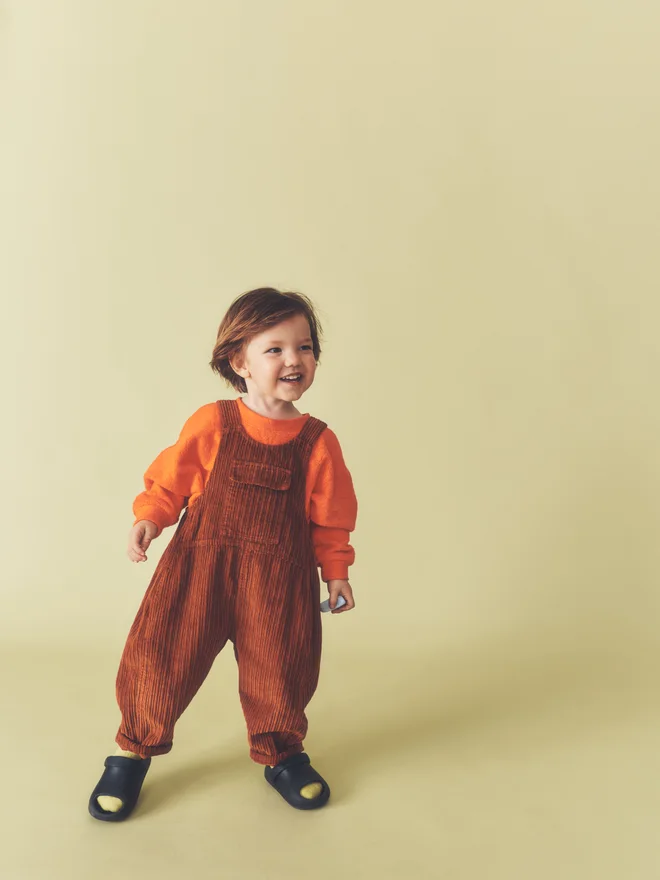 Toddler smiles mid-step against a pale yellow backdrop, wearing Studio Koter rust corduroy Kai dungarees over the Riverson top in Teddy Orange.