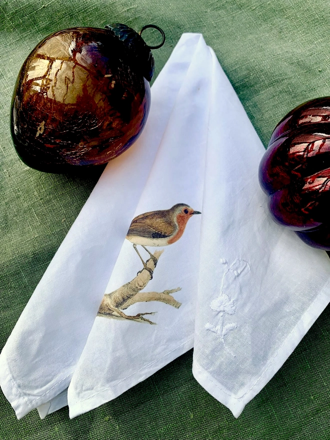 A white cotton napkin printed with a vintage illustration of a robin, alongside some large glass Christmas baubles