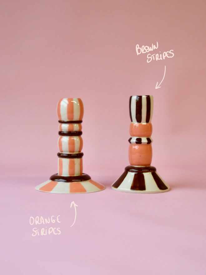 Handmade ceramic candlesticks with orange and brown stripe patterns, including an orange striped design on the left and a warm brown striped candlestick on the right, styled on a pink background.