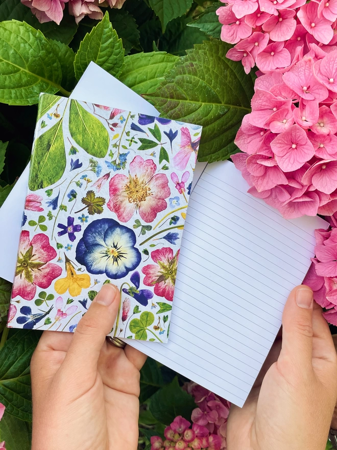 Hands Holding Two Small Botanical Print Notebooks with Pretty Pressed Flower Design on Pink Hydrangea Background