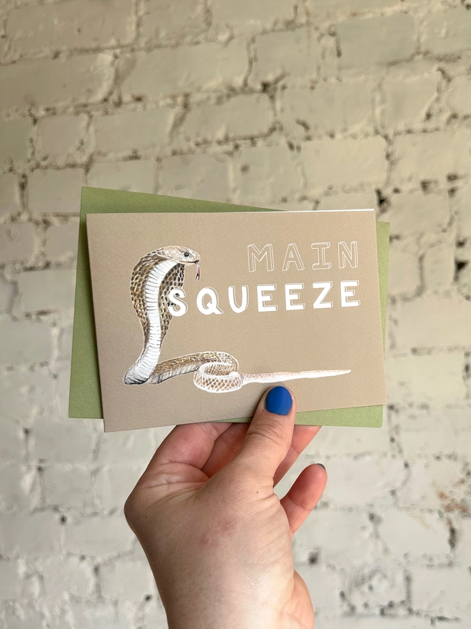 a beige greetings card featuring an illustration of a cobra with the text “main squeeze” with a green envelope