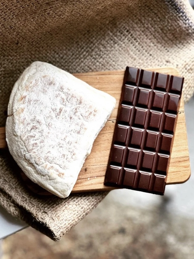 Irish Soda Bread Chocolate Bar, a chocolate bar unwrapped sitting on a wooden serving board. It is accompanied by some other food onna fabric surface. 