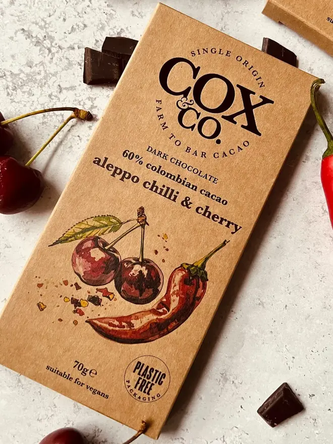 Aleppo Chilli & Cherry Chocolate Bar (Set Of 1, 2 Or 3), a chocolate bar sitting on a table surface. It is surrounded by small chocolate bits. 