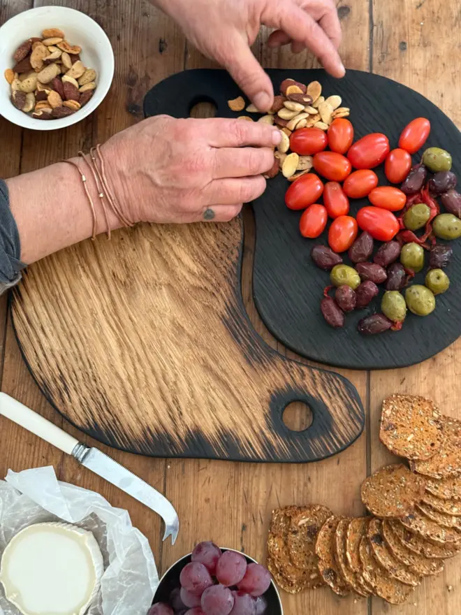 Hands styling the hug boards “Pair of handmade Yaki Sugi serving boards in blackened and natural oak, styled together with olives, cherry tomatoes, grapes, crackers and cheese on a rustic wooden table.”