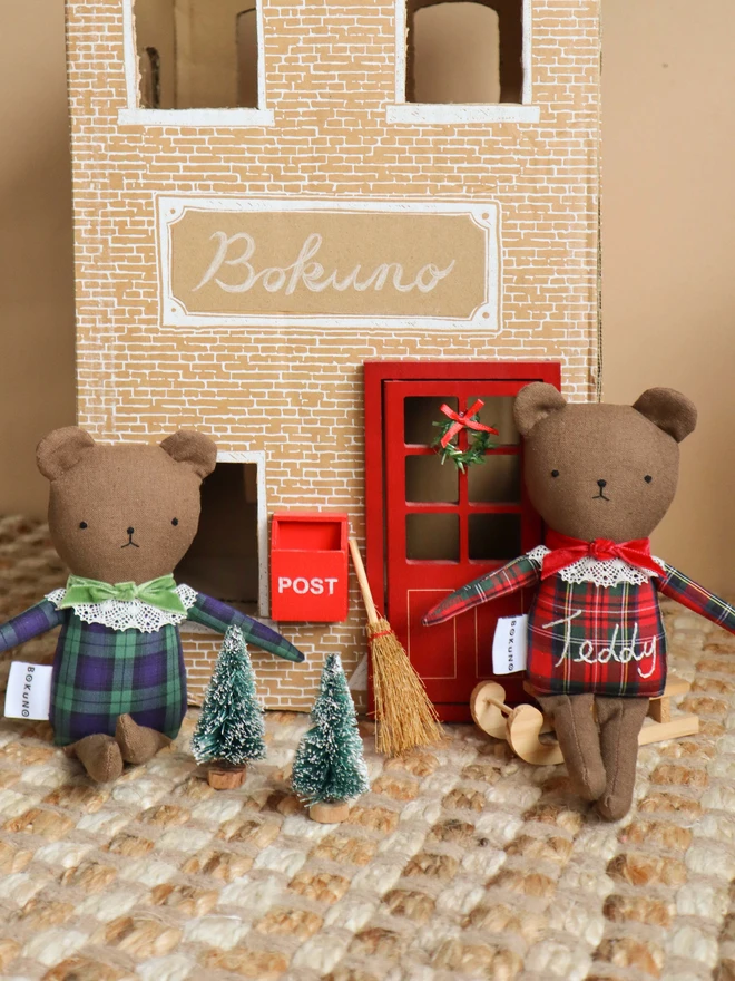 Two handmade linen teddy bears wearing festive tartan outfits, one in red and green with embroidered “Teddy” lettering and a red bow, and the other in green and navy tartan with a green bow. Both feature delicate lace collars and embroidered faces.
