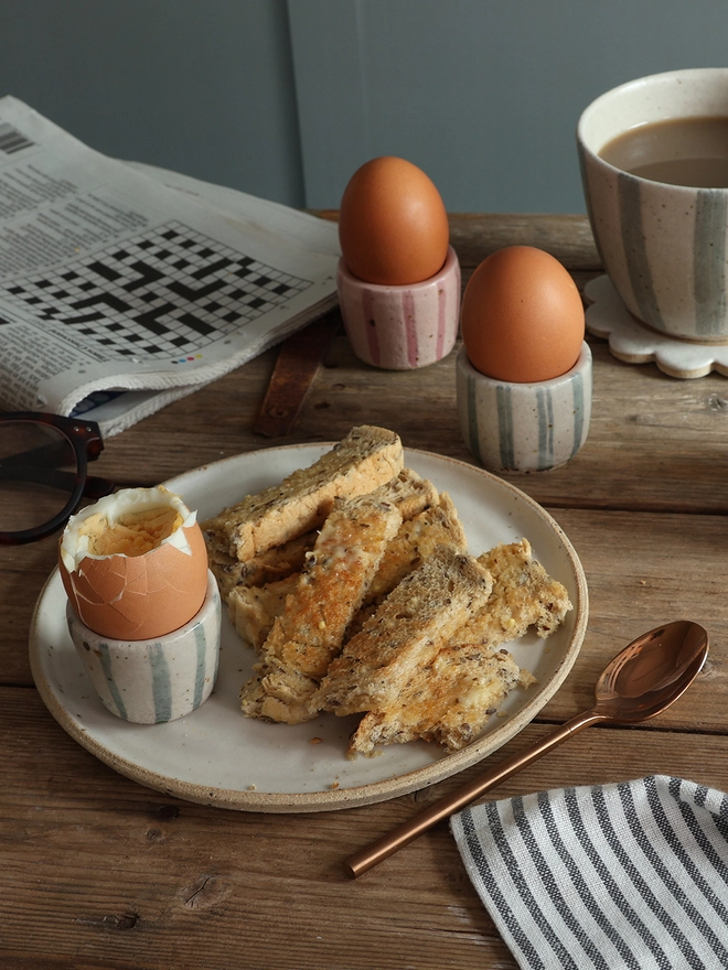 Two handmade striped ceramic egg cups with boiled eggs, toast, coffee and a newspaper on a breakfast table.