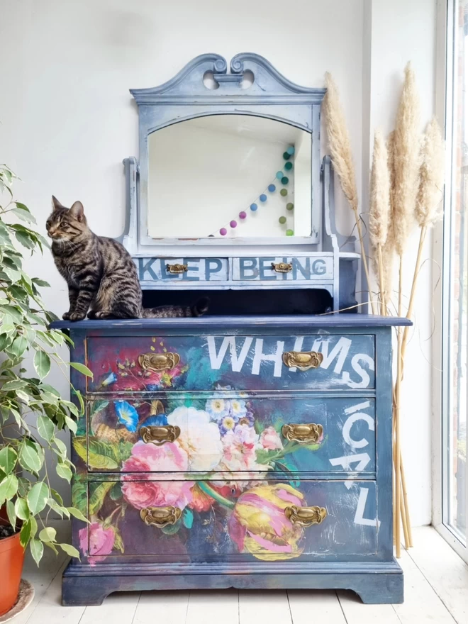 Painted vintage table in dark and light blue with a large, statement decoupage floral design and the wording ‘keep being whimsical’ across the front. 