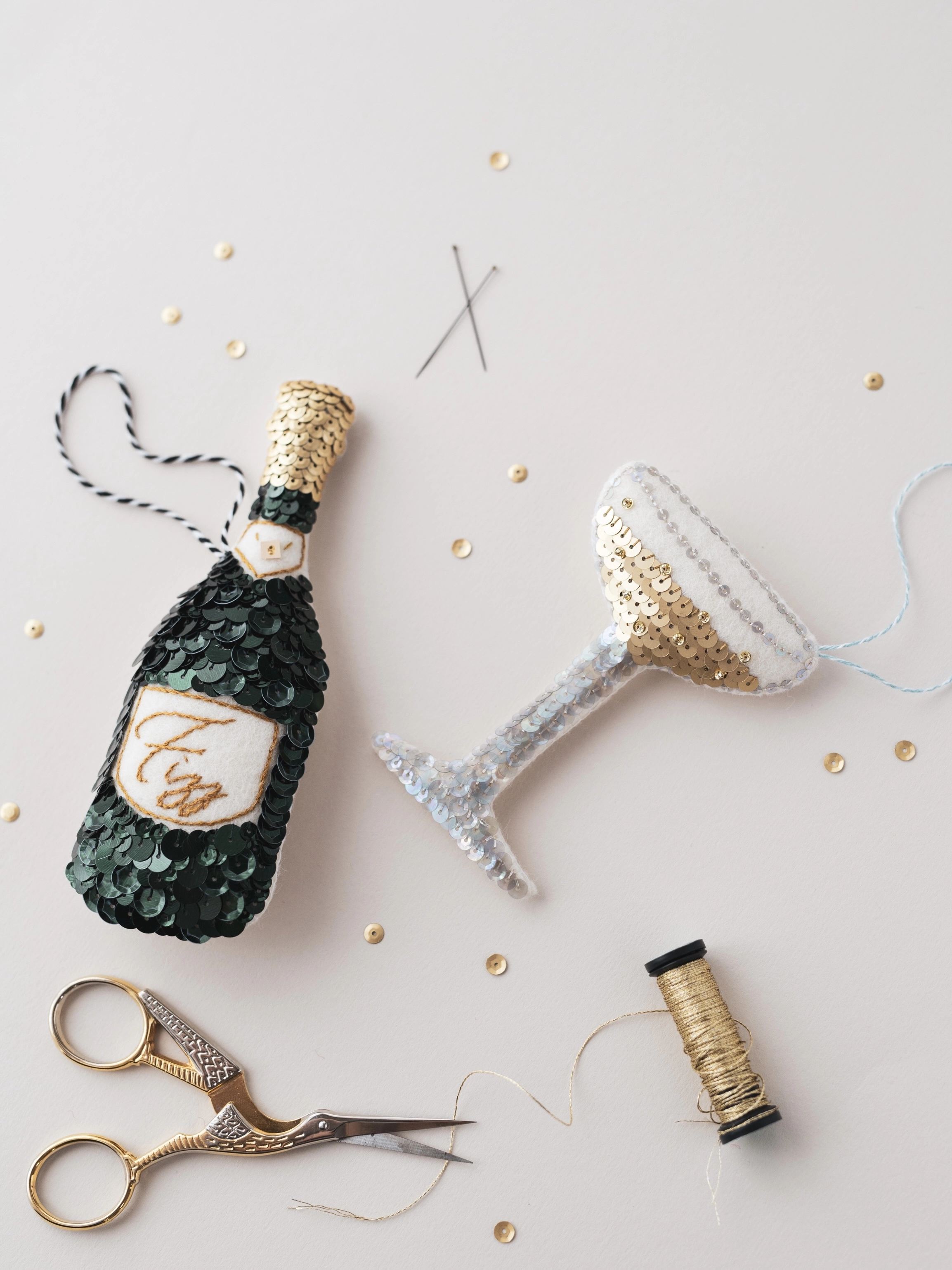 a sequinned green champagne bottle ornament and golden sequinned champagne coupe glass ornament on a white table