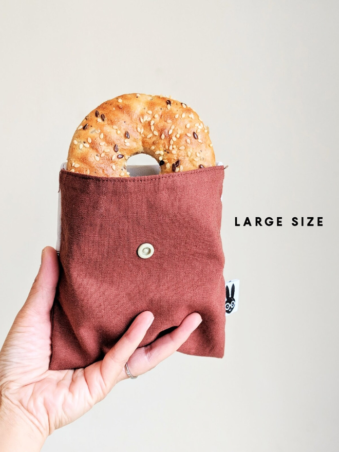 reusable linen snack pouch. a rust brown pouch holding a bagel.