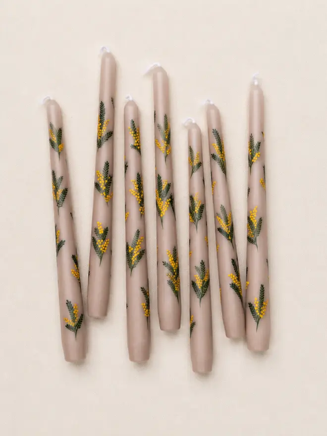 Eight hand-painted oat-coloured taper candles with a yellow mimosa floral design, arranged at varying angles on a textured linen background.