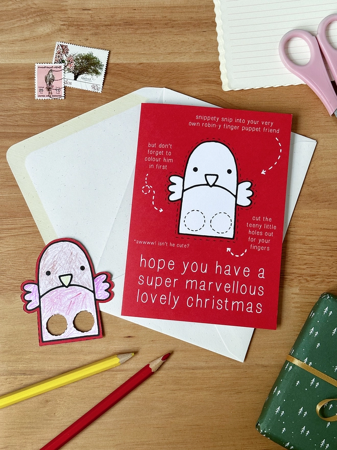 A colour in Christmas card with a robin finger puppet design lays on a white envelope on a wooden desk.