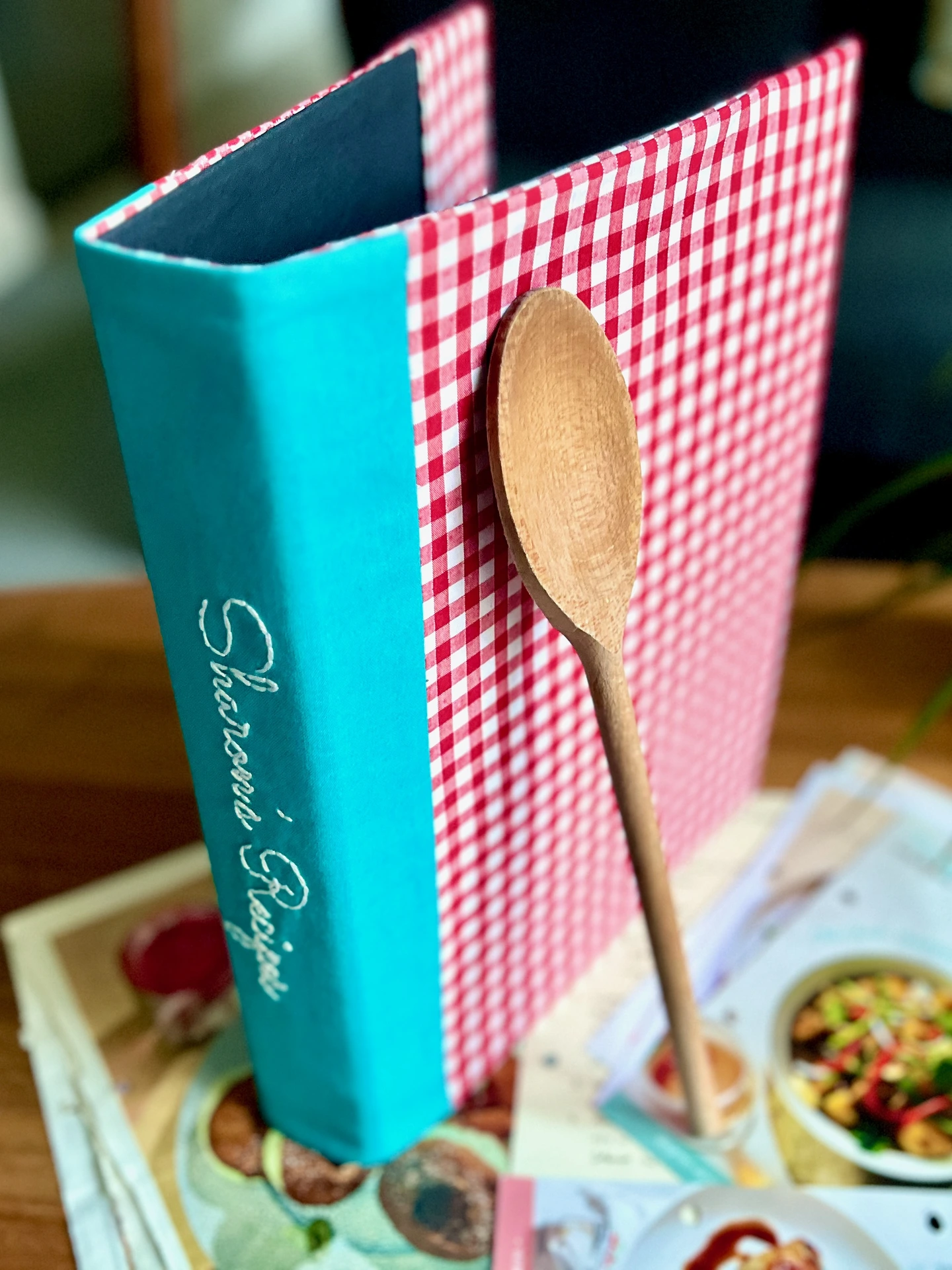 Red gingham binder with personalisation embroidered on teal spine, standing on wooden table.