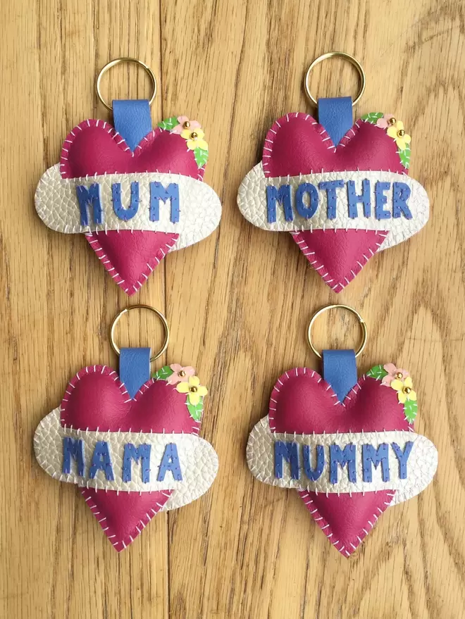 Four pink heart keyrings on a wooden surface. One spells MUM in blue lettering on a white scroll, the others spell MUMMY, MUM and MOTHER