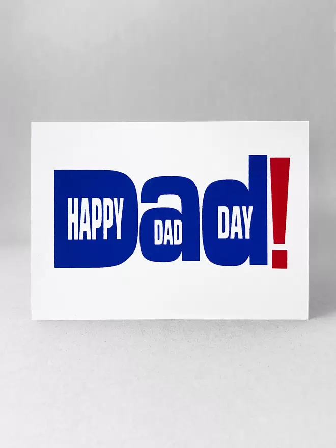 Studio shot of white landscape greetings card on light grey background, flat on facing the front. The design is typography of the word Dad! With happy Dad Day written within the letters. The writing is printed in royal blue with an oversized exclamation mark in red to the right. 