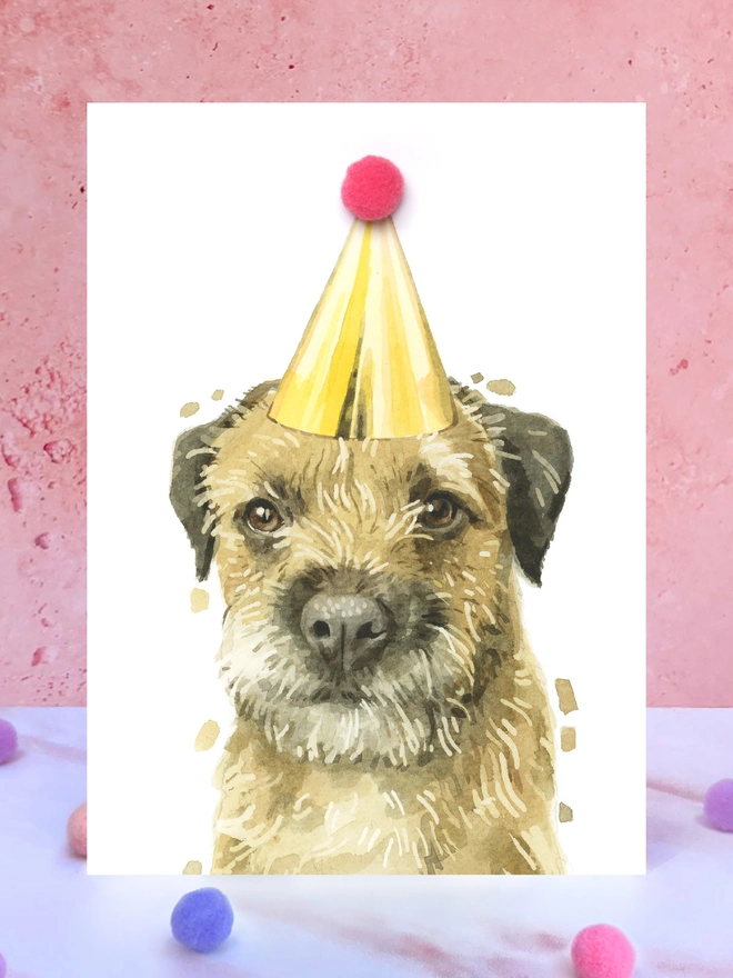 A greeting card featuring a hand painted design of a border terrier wearing a pompom adorned party hat. The hat is stood up right on a marble surface surrounded by pompoms.