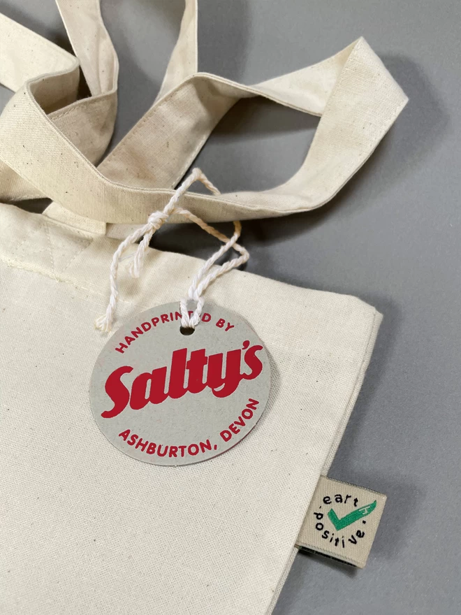 Close up of a white, natural coloured tote bag laid down with the  Saltys swing tag tied to one strap and the earthpositive tag showing sewn into the seam.