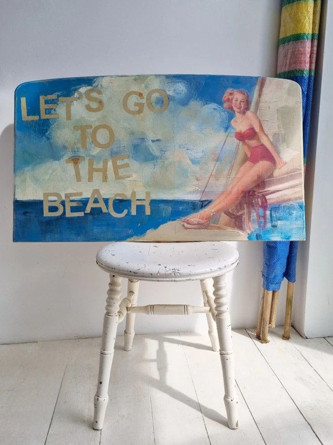 Upcycled art panel in blues and creams featuring a seascape and an image of a retro pin-up lady on a boat and the text reads ‘let’s go to the beach’ in a distressed, shabby chic style