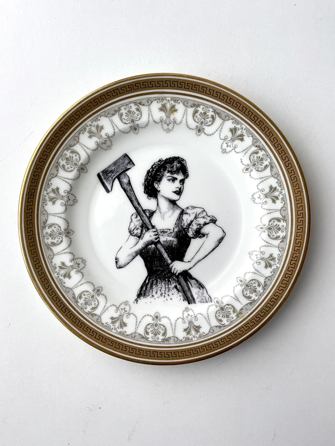 a vintage plate with a gold ornate border, in the centre is a black and white vintage image of a victorian pretty lady wielding a huge axe