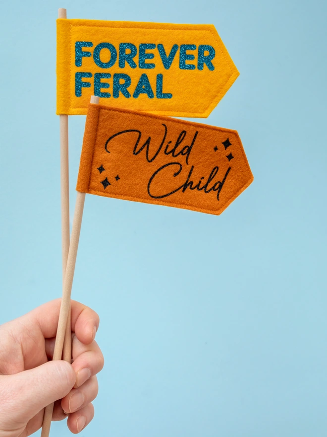 2 handheld felt flags, 1 that has the words "forever feral" written on it and the other "wild child"