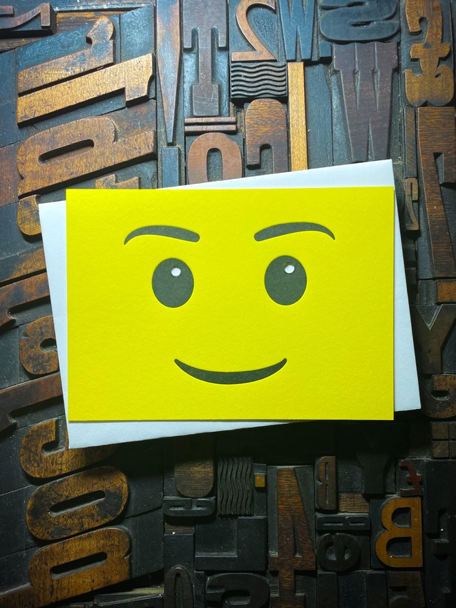 A bright yellow letterpress card with a smiling face emoji is placed on top of a white envelope. The background consists of various wooden printing letters in different orientations and sizes.