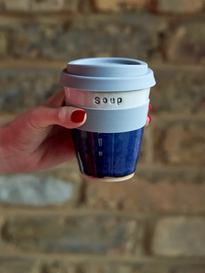 Ceramic midnight blue and white travel mug with a silicon band and lid, stamped with the word 'soup', held up against a brick background