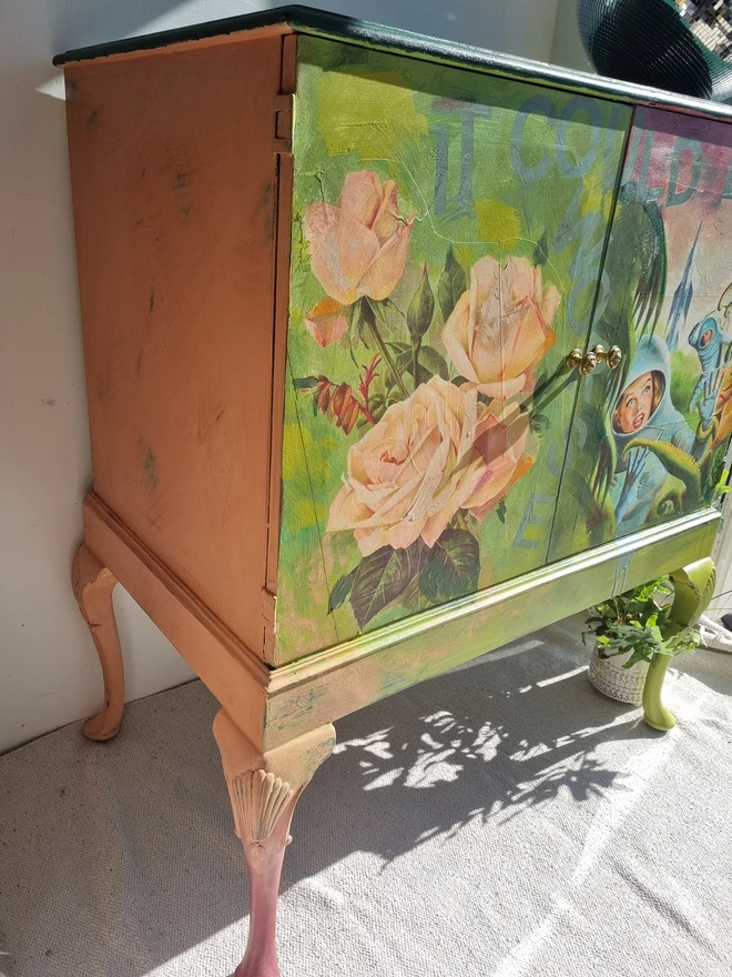 A painted record cabinet decorated with a sci fi image from the 1950s of a lady running from an alien and on the other door, a rose taken from a Victorian seed packet. There is wording that runs down the middle of the cabinet which reads 'it could be worse'. The colours are apple green and pinks.