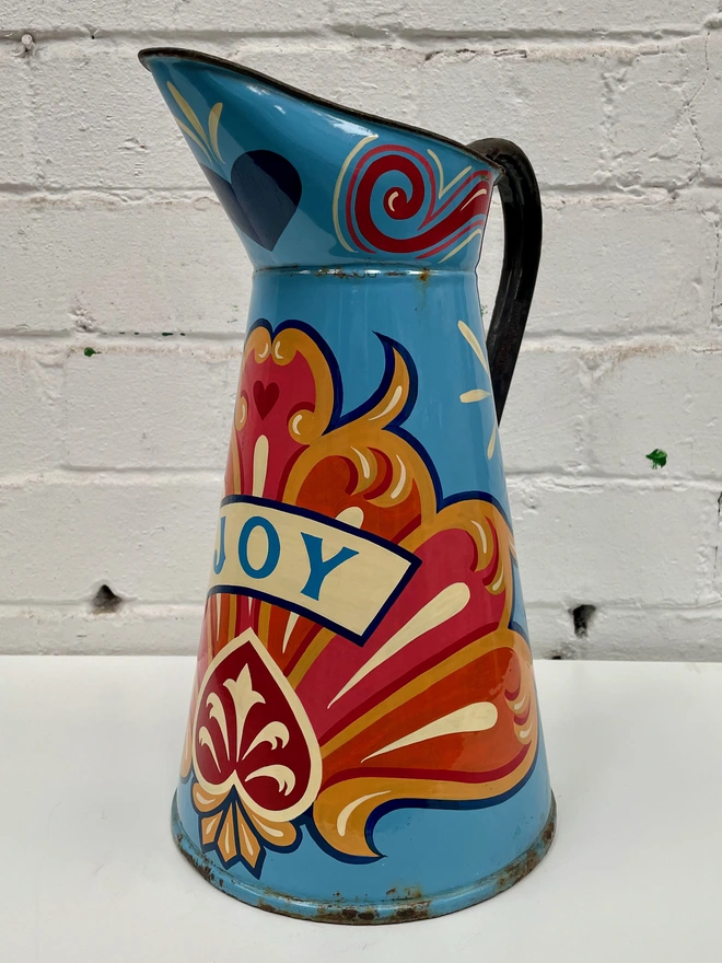 Large blue enamel jug hand-painted with a traditional fairground inspired design, in pink, orange and yellow. 'Joy' is painted in a banner across the centre.