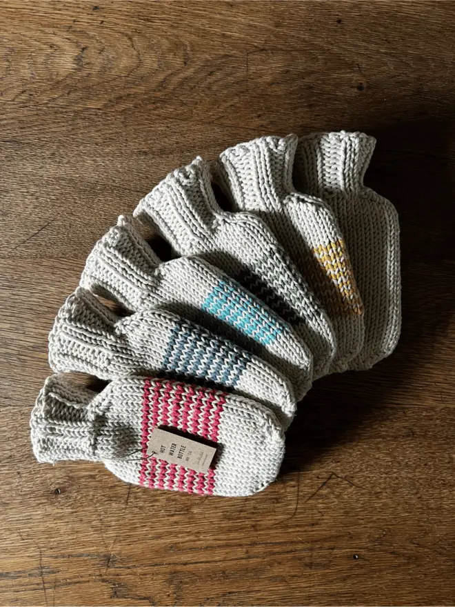 Six mini hot water bottles with striped knitted cotton string cover fanned out on an oak table