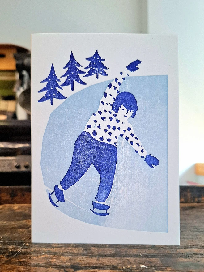 A handprinted greetings card standing open on a wooden surface.  The card has a handprinted illustration on it, showing a smiling girl iceskating.  It is printed in light blue and navy.