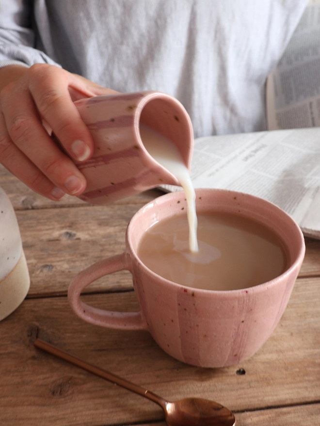 A female hand holding a Handmade Ceramic Stoneware Small Jug in Pink Stripe, pouring milk from the jug into a mug in the same style filled with coffee. A brass spoon, a newspaper and a ceramic sugar pot sit around the mug. 