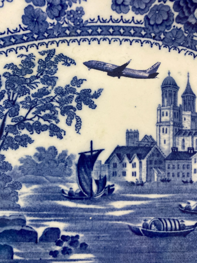 Classic blue and white patterned plate featuring a castle and river with boats sailing along. A Jet2 plane flies high in the sky.