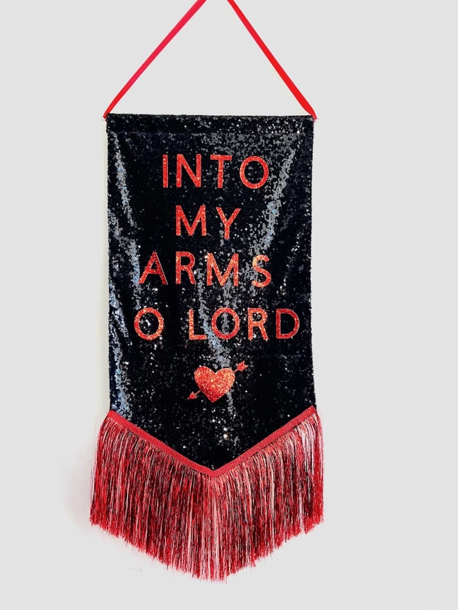 A black sequin banner with 'INTO MY ARMS O LORD' and an image of a heart features in red. It hangs on red ribbon and features a red tinsel trim along the bottom.