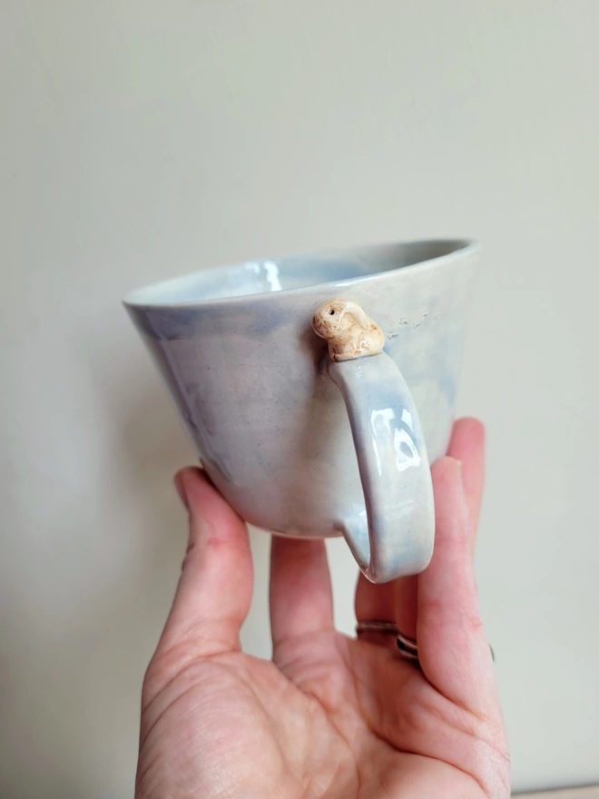 Grey hand-made cup with miniature beige bunny rabbit and pawprints held in a hand