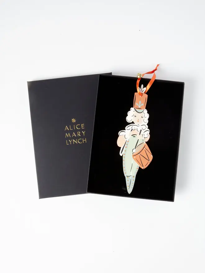 Little Drummer Tree Dancer. wooden hanging little drummer decoration in an open black gift box embossed "alice mary lynch" in gold, with a curly white wig, terracotta hat and drum, sage green uniform, orange ribbon with a jingle bell