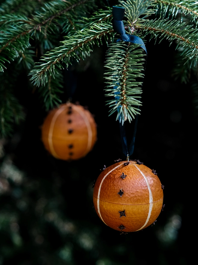 Handcrafted orange and clove scented pomander bauble hanging from a fir Christmas tree, surrounded by soft green foliage and warm natural tones.